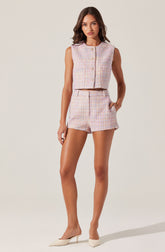 A woman is wearing a sleeveless, pastel-colored, textured knit crop top with a round neckline and button closure at the front. She pairs it with matching high-waisted shorts that have a similar textured knit fabric and a notched hem. She completes the outfit with pointed-toe, cream-colored high heels.